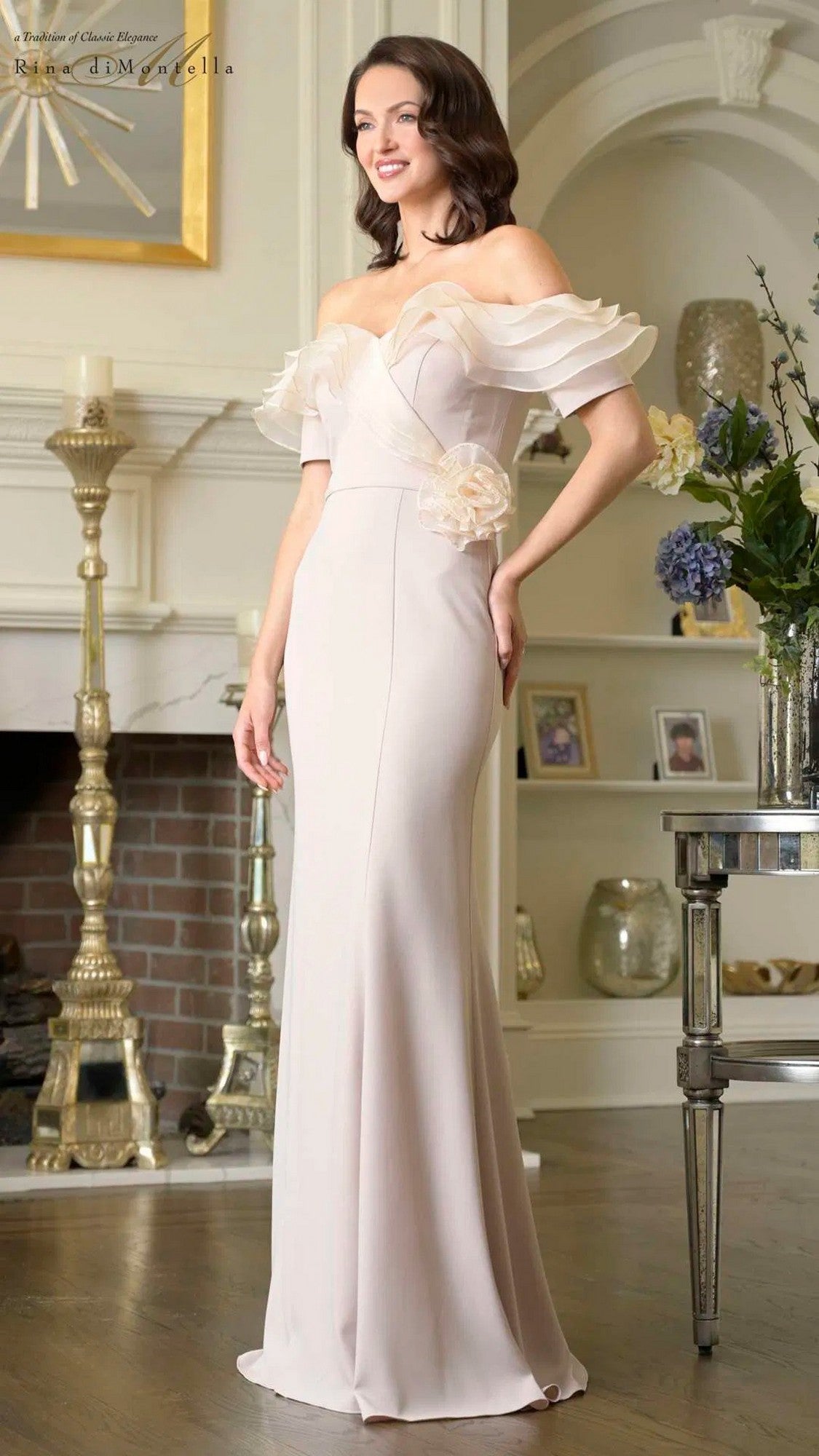 A woman stands indoors wearing the Rina Di Montella RD3418 long formal dress in champagne, featuring an off-the-shoulder neckline, ruffled sleeves, and a floral accent at the waist. A fireplace and decor are seen in the background.